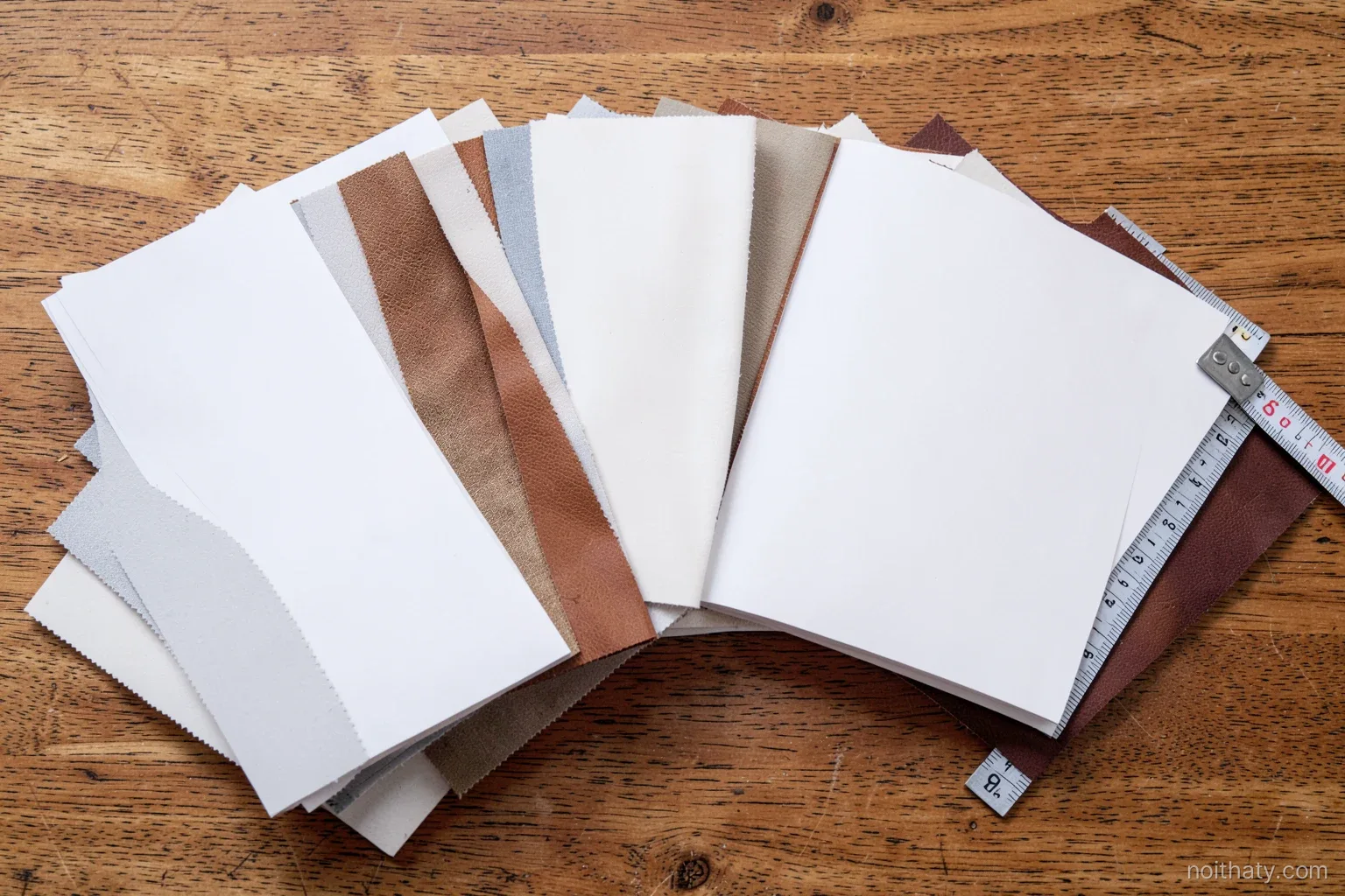 Top-down view of fabric and leather swatch books fanned open beside wood veneer samples and a tape measure on an oak table, blank pages, soft natural light