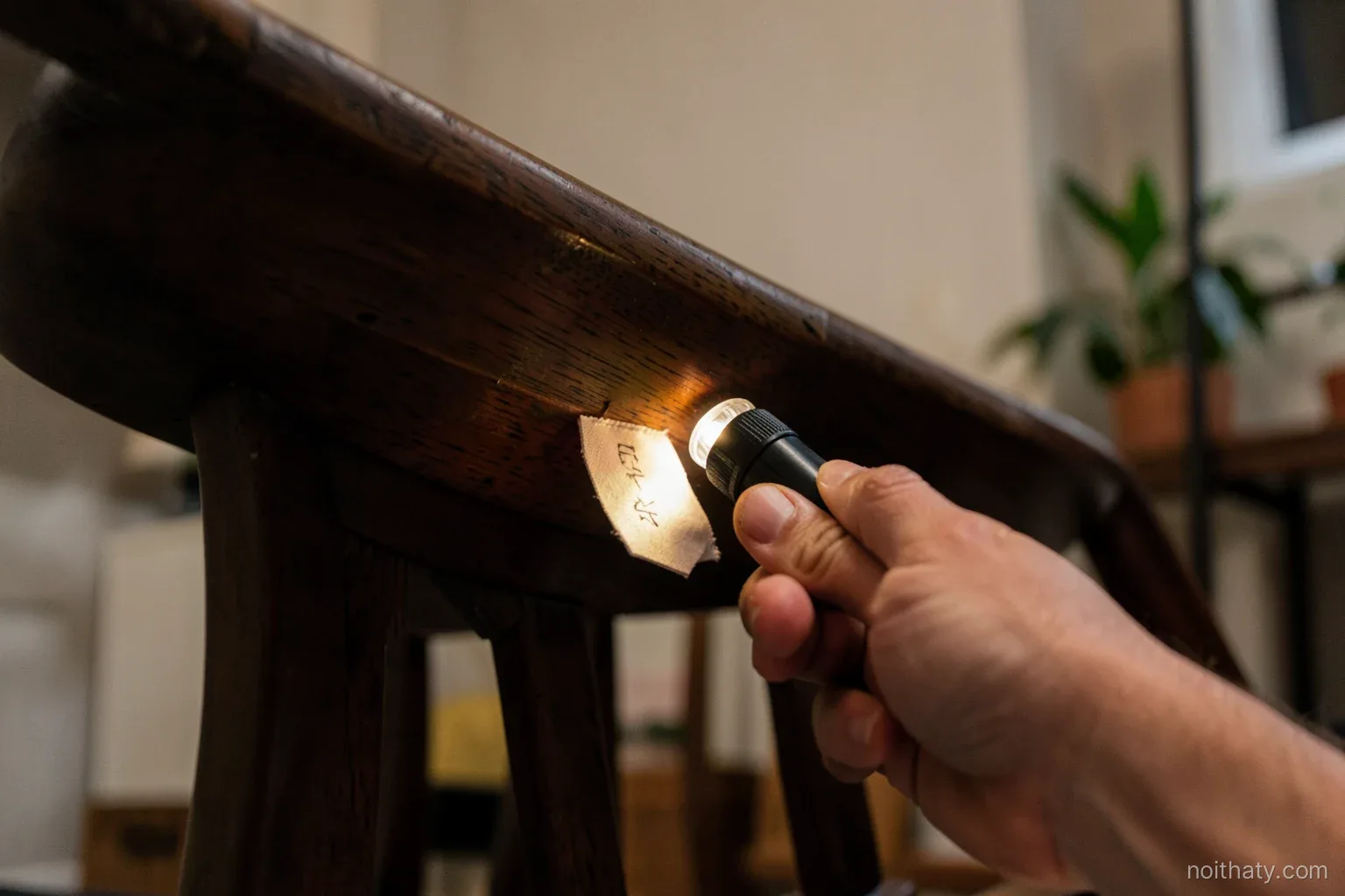 Low angle close-up of a hand with a small flashlight examining the underside of an upturned wooden chair, a blank fabric tag visible without writing, warm lamp light, dark walnut